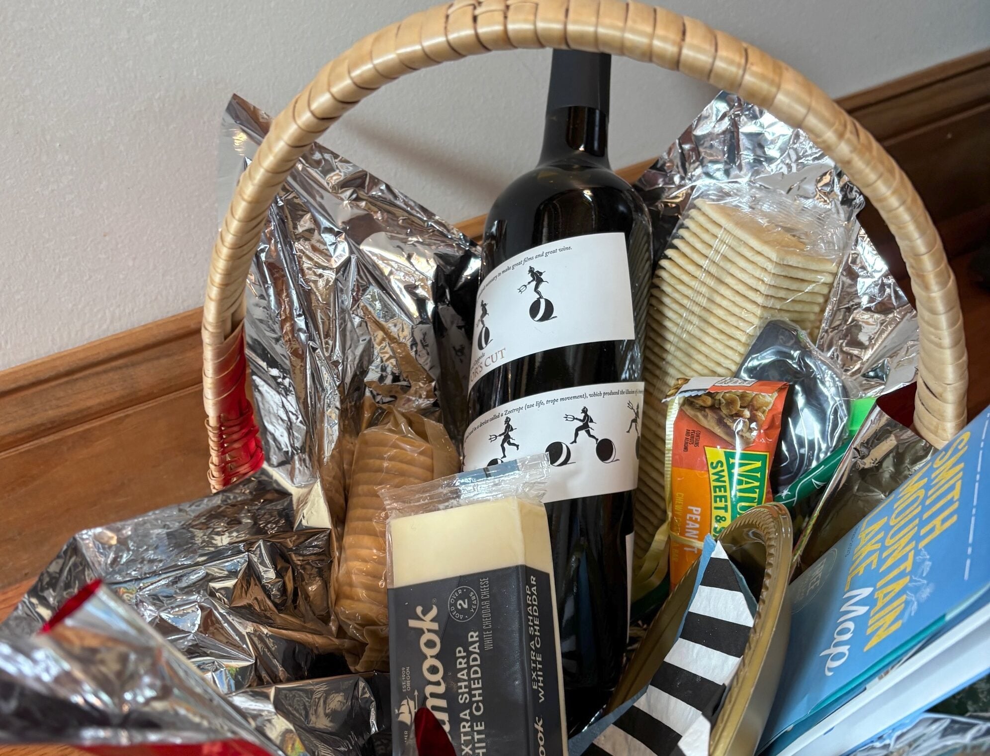 Gift basket containing a wine bottle, crackers, and cheese wrapped in foil on a wooden surface