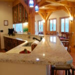 Open-plan kitchen with a curved granite island, wooden cabinets, and exposed timber beams; dining area beyond with large windows.