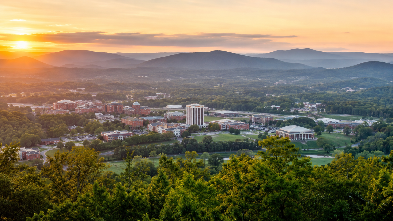 Sunset over a college town with brick buildings, a central tall tower, and surrounding green hills and trees