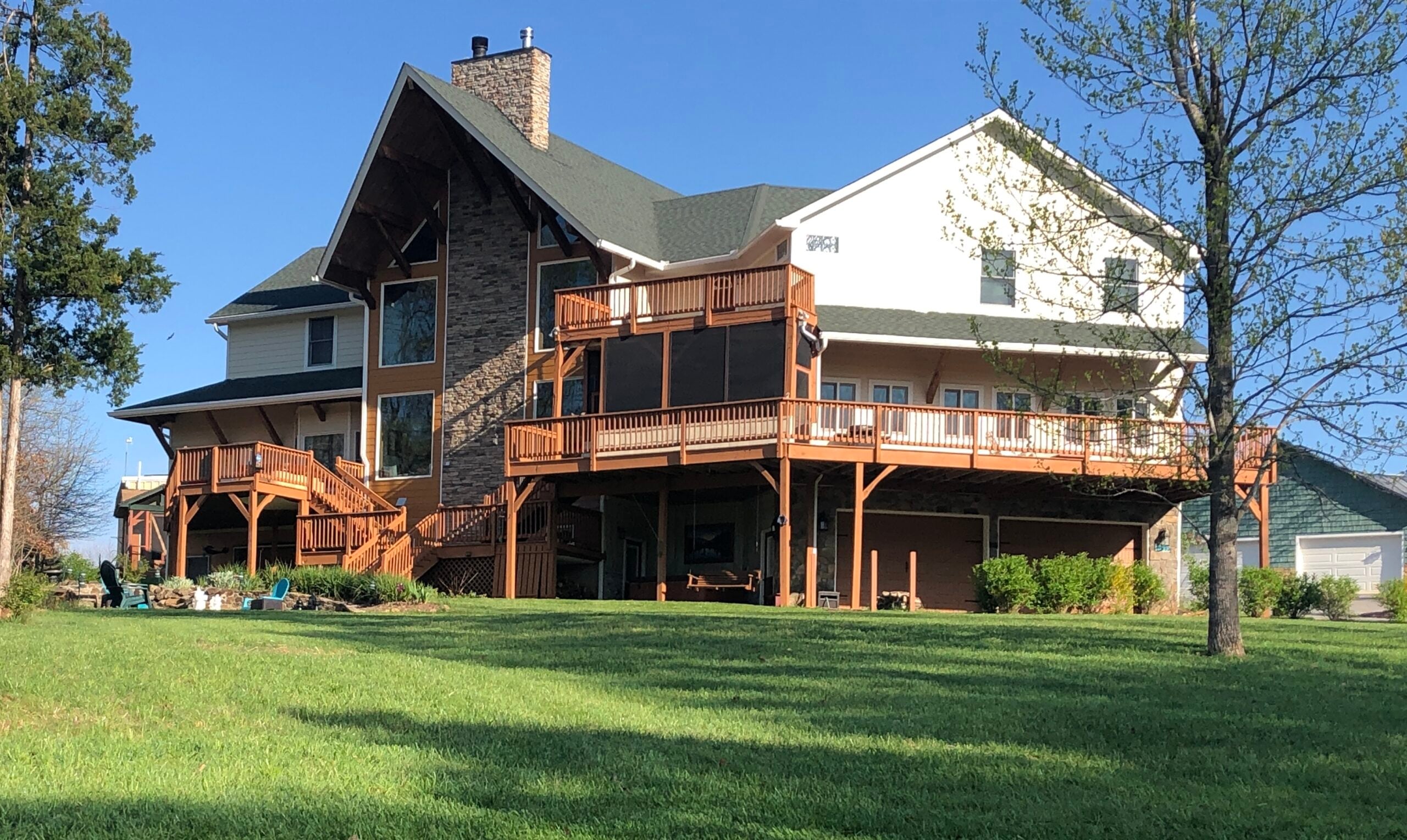 Two-story house with wraparound wooden decks, stone chimney, and green roof, set against a blue sky and trees with a manicured lawn below.