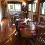 Wooden dining room with a long table set for a meal, colorful placemats, and tall red candles under exposed beam ceilings.