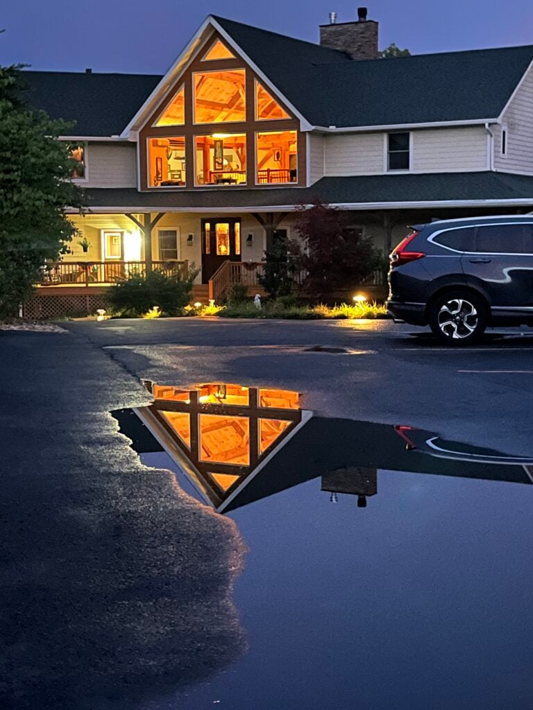 A two-story house at dusk with large illuminated triangular windows, warm interior light visible through the front windows and porch area, reflected in a puddle in the foreground.