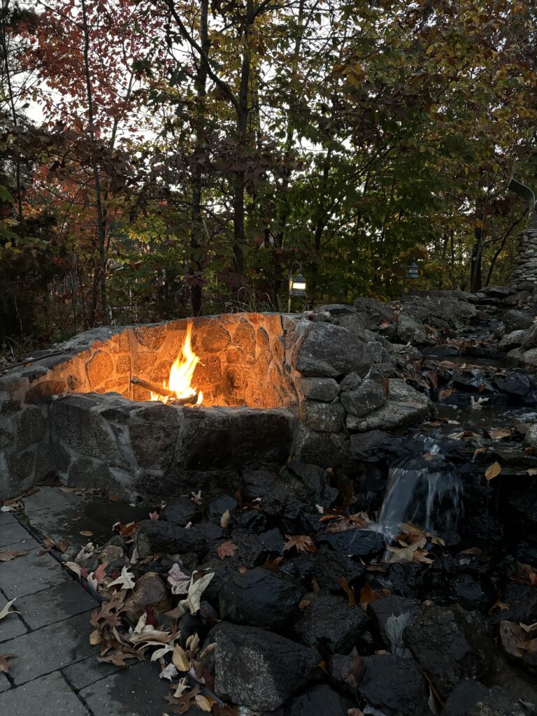 Stone fire pit with a bright flame, surrounded by rocks and fallen autumn leaves, with trees in fall colors behind it.