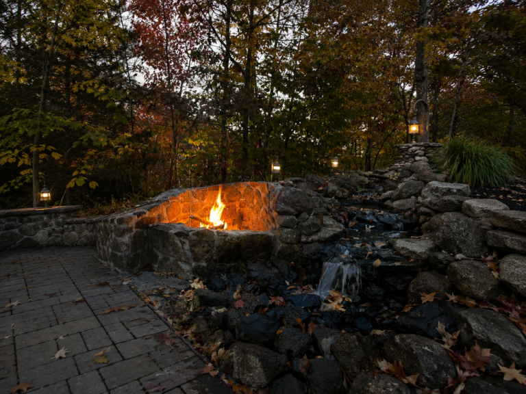 Stone fire pit surrounded by a rock wall, with bright flames, lanterns along the path, and autumn trees in the background at dusk.