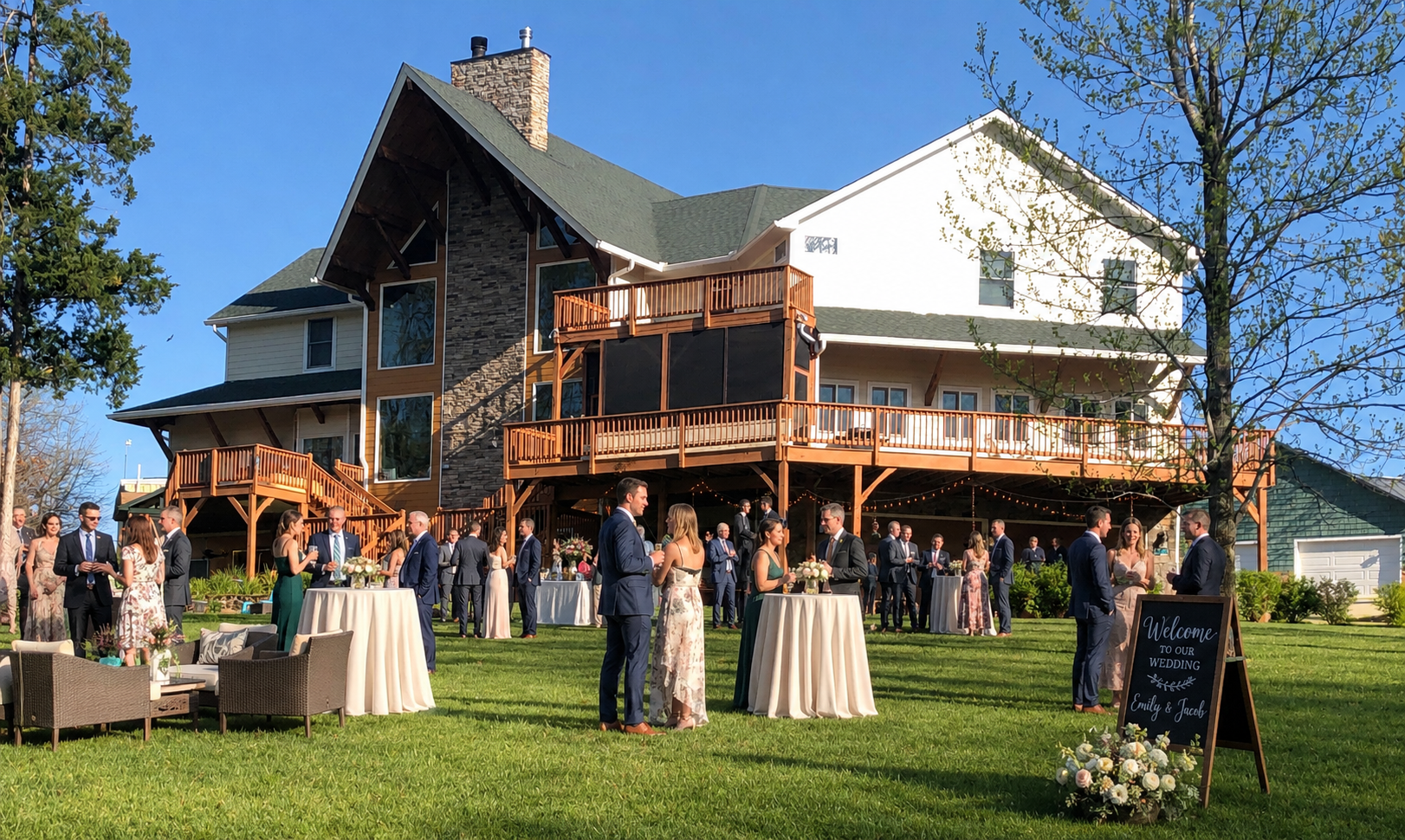 Outdoor wedding reception on a sunny lawn in front of a large wooden-clad house; guests mingle around high-top tables and string lights.