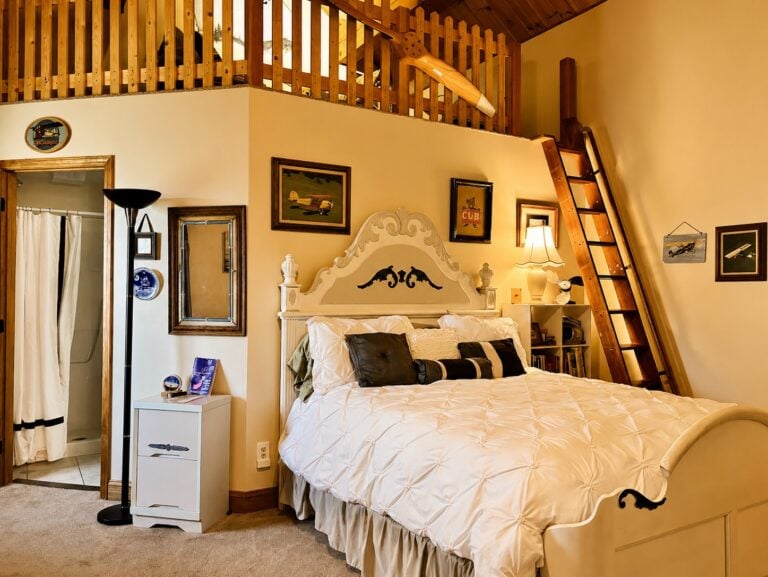 Aviation-themed loft guest room at Bedford Landings Bed & Breakfast near Smith Mountain Lake, featuring a cozy white bed, wood-paneled loft, ladder, private bathroom, and warm cabin-style decor.