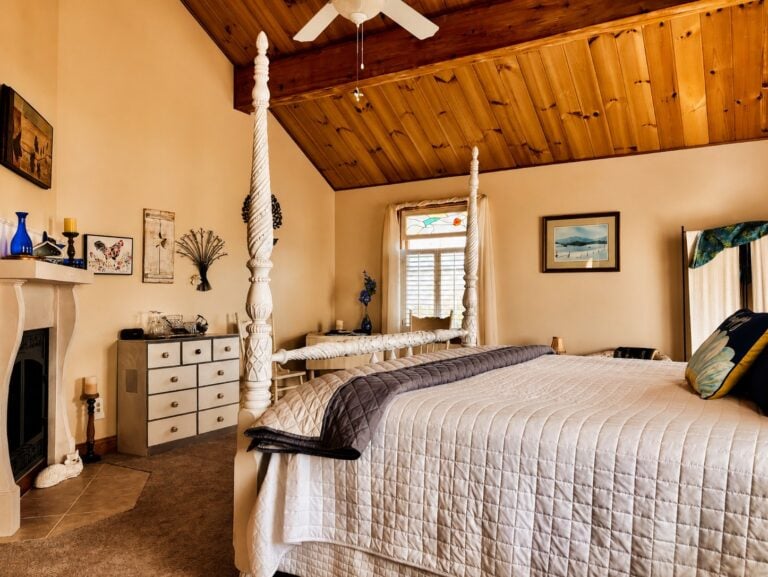 Guest room at Bedford Landings Bed & Breakfast near Smith Mountain Lake, featuring a white four-poster bed, vaulted wood ceiling, cozy fireplace, stained-glass window, and warm natural light.
