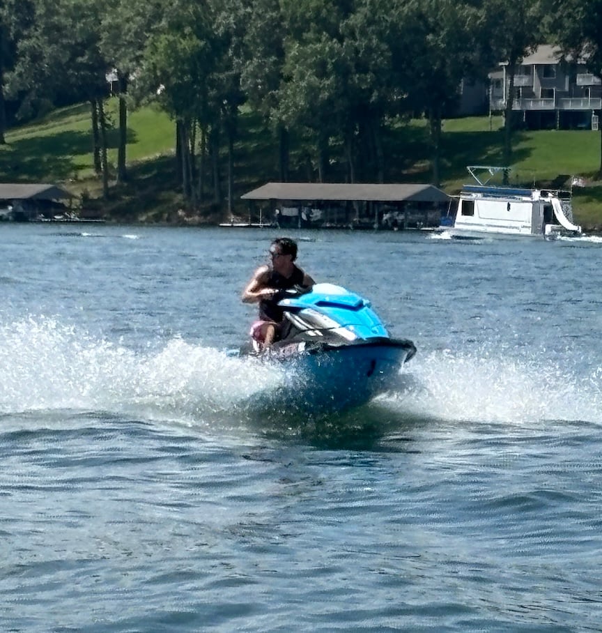 Person riding a blue jet ski at speed on a lake, spraying water with a tree-lined shore in the background.