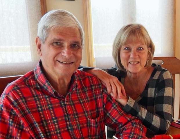 Smiling older couple posing together indoors; man in red plaid shirt, woman resting her hand on his shoulder from behind.