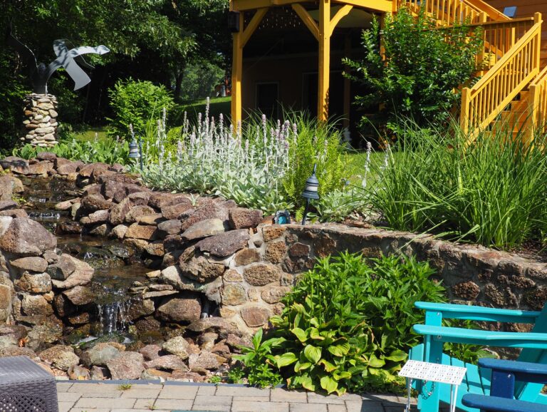 Rock-lined waterfall feature in a landscaped backyard with tall grasses, lavender plants, and a yellow wooden deck in the background.