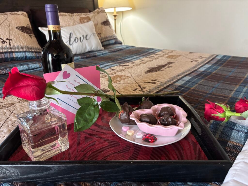 Romantic bedside tray on a quilted bed: a bottle of wine, pink rose, heart card, and chocolate candies.